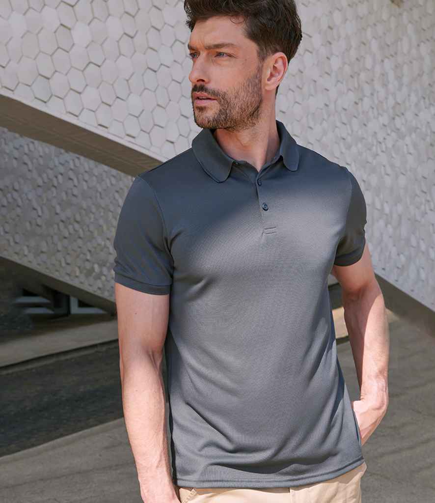 A man with short brown hair and a beard wears a Henbury Recycled Polyester Piqué Polo Shirt in dark gray and beige pants, standing outdoors before a hexagonal tiled wall. He looks left with one hand in his pocket. Brand: Henbury.