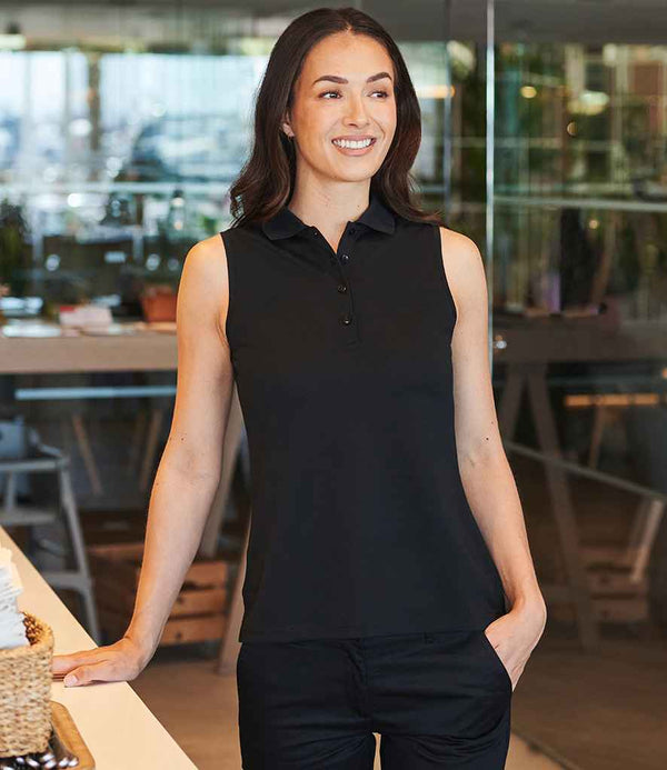 A woman wears a Henbury Ladies Sleeveless Coolplus® Polo Shirt and black pants indoors, smiling as she rests one hand on a counter. Behind her are tables, chairs, and glass walls.