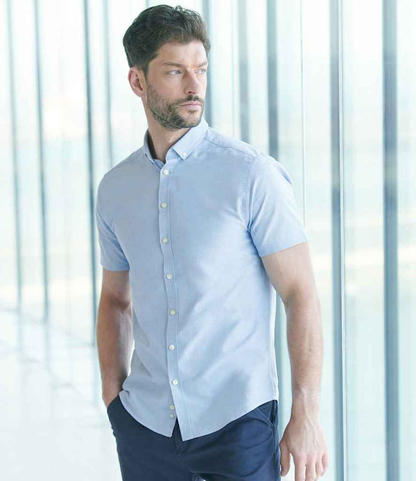 A man with short brown hair and a beard is wearing the Henbury Modern Short Sleeve Slim Fit Oxford Shirt by Henbury in light blue with dark pants, standing indoors near large glass windows and looking to his left.
