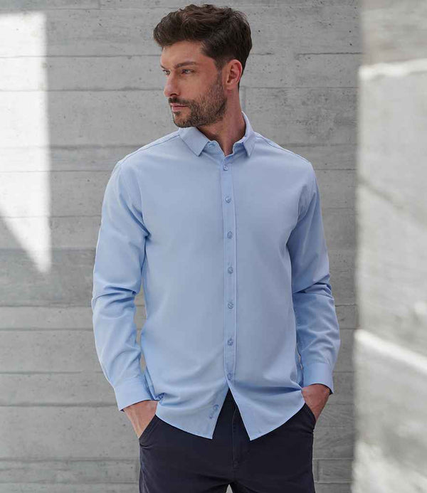 A man wearing a Henbury Long Sleeved Cotton Feel Coolplus® Shirt in light blue, featuring a button down collar, and dark pants stands against a concrete wall, facing left with his hands in his pockets.