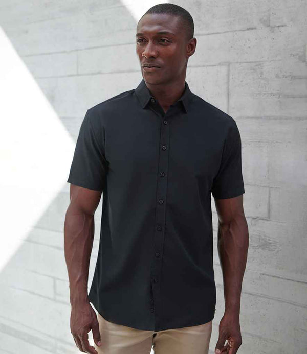 A man stands against a light concrete wall, wearing the Henbury Short Sleeved Cotton Feel Coolplus® Shirt in black with beige pants. He looks to the side with a neutral expression.