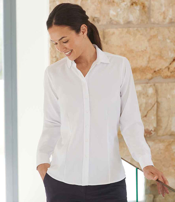 A woman with dark hair tied back wears the Henbury Ladies Long Sleeve Wicking Shirt by Henbury with dark pants, standing indoors against a stone wall, hands in her pockets, looking down and smiling.
