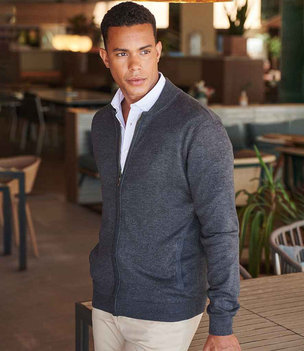 A man wears the Henbury Unisex Zip Through Cardigan by Henbury—dark gray, with ribbed cuffs and a baseball collar—over a white shirt and beige pants, standing indoors by a wooden table amid chairs, looking to his left.