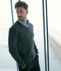 A man with dark hair and a beard stands indoors by large windows overlooking a waterfront, wearing the Henbury Lightweight Cotton Acrylic V Neck Sweater in dark gray over a light blue collared shirt, one hand in his pocket, looking thoughtfully to the side.