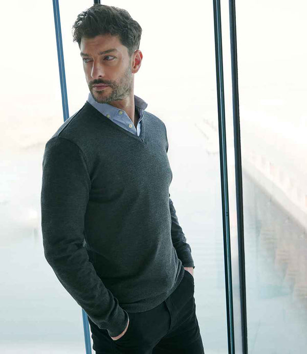 A man with dark hair and a beard stands indoors by large windows overlooking a waterfront, wearing the Henbury Lightweight Cotton Acrylic V Neck Sweater in dark gray over a light blue collared shirt, one hand in his pocket, looking thoughtfully to the side.