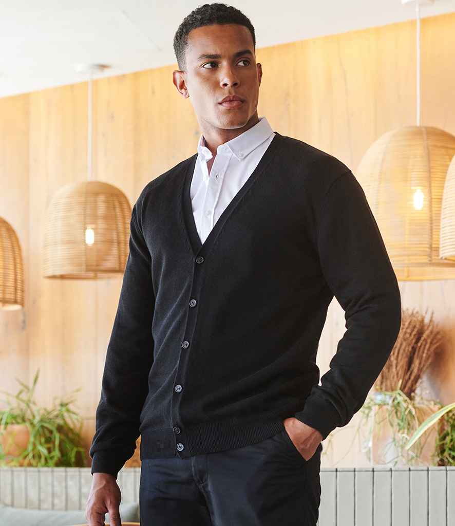 A man stands indoors in a modern room, wearing the Henbury Lightweight Cotton Acrylic V Neck Cardigan—a black cardigan with horn-effect buttons—over a white shirt.