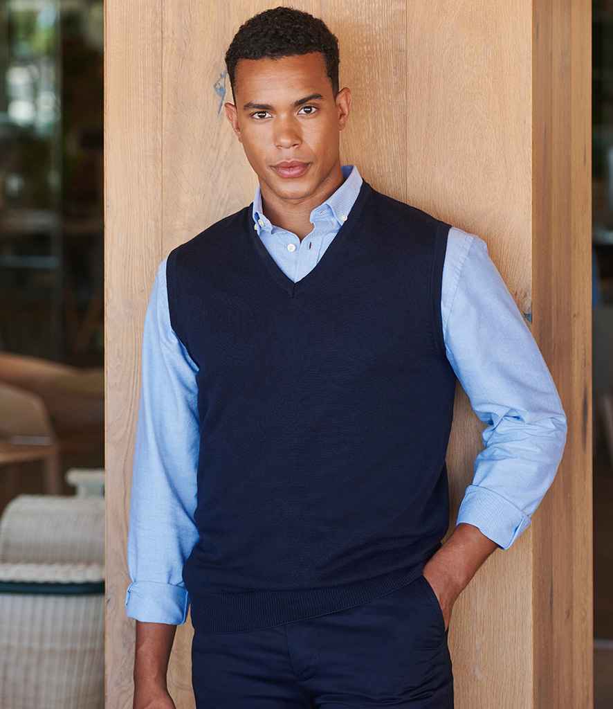 A man stands indoors in front of a wooden wall, wearing the Henbury Lightweight Sleeveless Cotton Acrylic V Neck Sweater over a light blue button-down shirt, with ribbed armholes and one hand in his pocket.