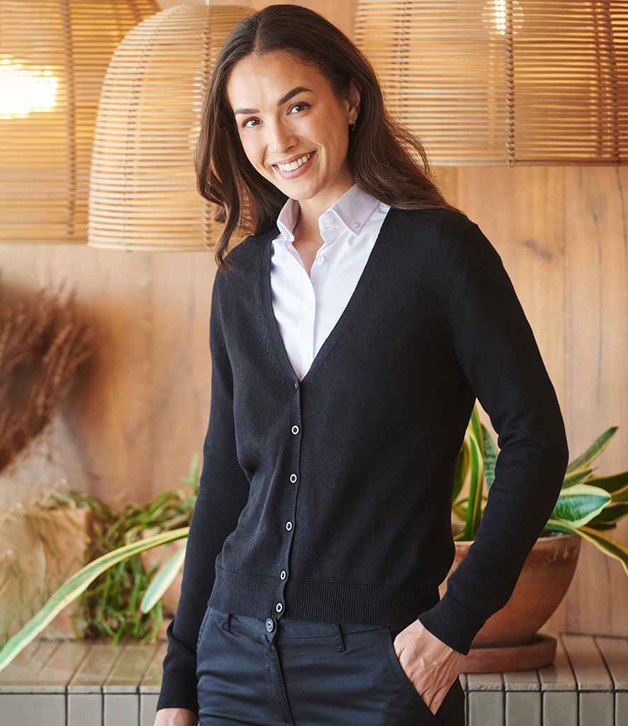 A woman with long brown hair is standing indoors, smiling, and wearing a white button-up shirt under the Henbury Ladies Lightweight V Neck Cardigan by Henbury. Plants and woven light fixtures can be seen in the background.