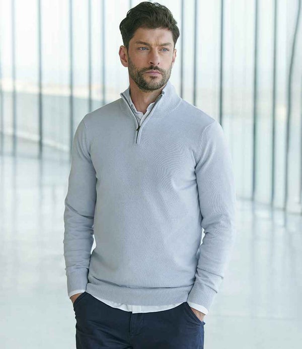 A man with short brown hair and a beard wears the Henbury Zip Neck Sweater by Henbury, a fine knit light gray quarter-zip with ribbed collar and metal zip, over a white shirt and dark pants, standing indoors before large glass windows.