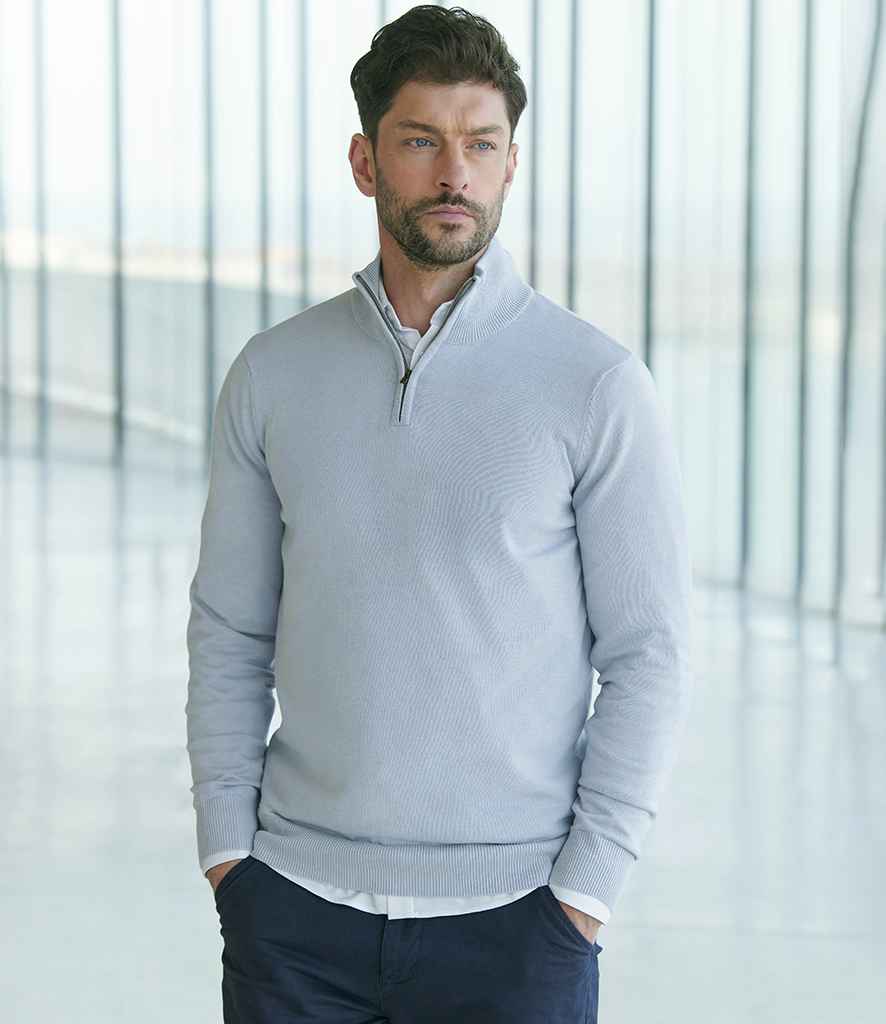 A man with short brown hair and a beard wears the Henbury Zip Neck Sweater by Henbury, a fine knit light gray quarter-zip with ribbed collar and metal zip, over a white shirt and dark pants, standing indoors before large glass windows.
