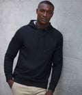 A person wearing a Henbury Unisex Sustainable Hoodie in black and light-colored pants stands against a light gray concrete wall, looking directly at the camera with a neutral expression.