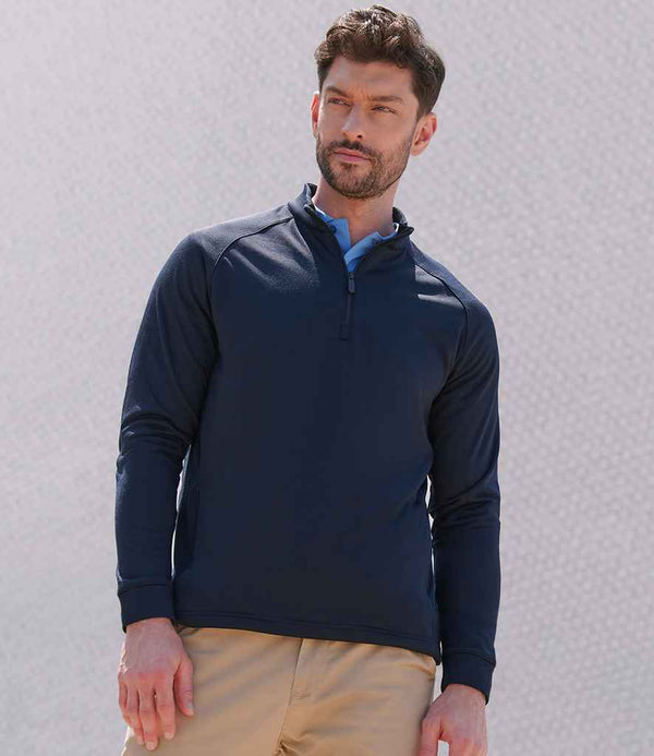 A man with short dark hair and a trimmed beard stands outdoors against a light textured background, wearing the Henbury Zip Neck Wicking Top in navy blue with raglan sleeves and khaki pants, looking slightly to the side.