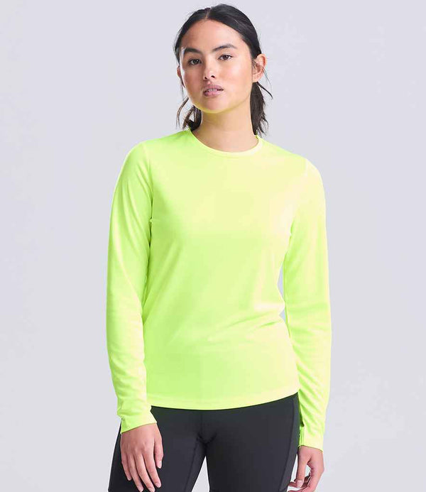 A person faces the camera with a neutral expression, wearing the AWDis Ladies Cool Long Sleeve T-Shirt by Just Cool in neon yellow and black pants against a plain background. The shirt is made from recycled polyester.