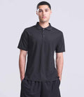 A person stands facing forward, wearing a Just Cool AWDis SuperCool™ Performance Polo Shirt in black with black shorts, hands in their pockets against a neutral light gray background.