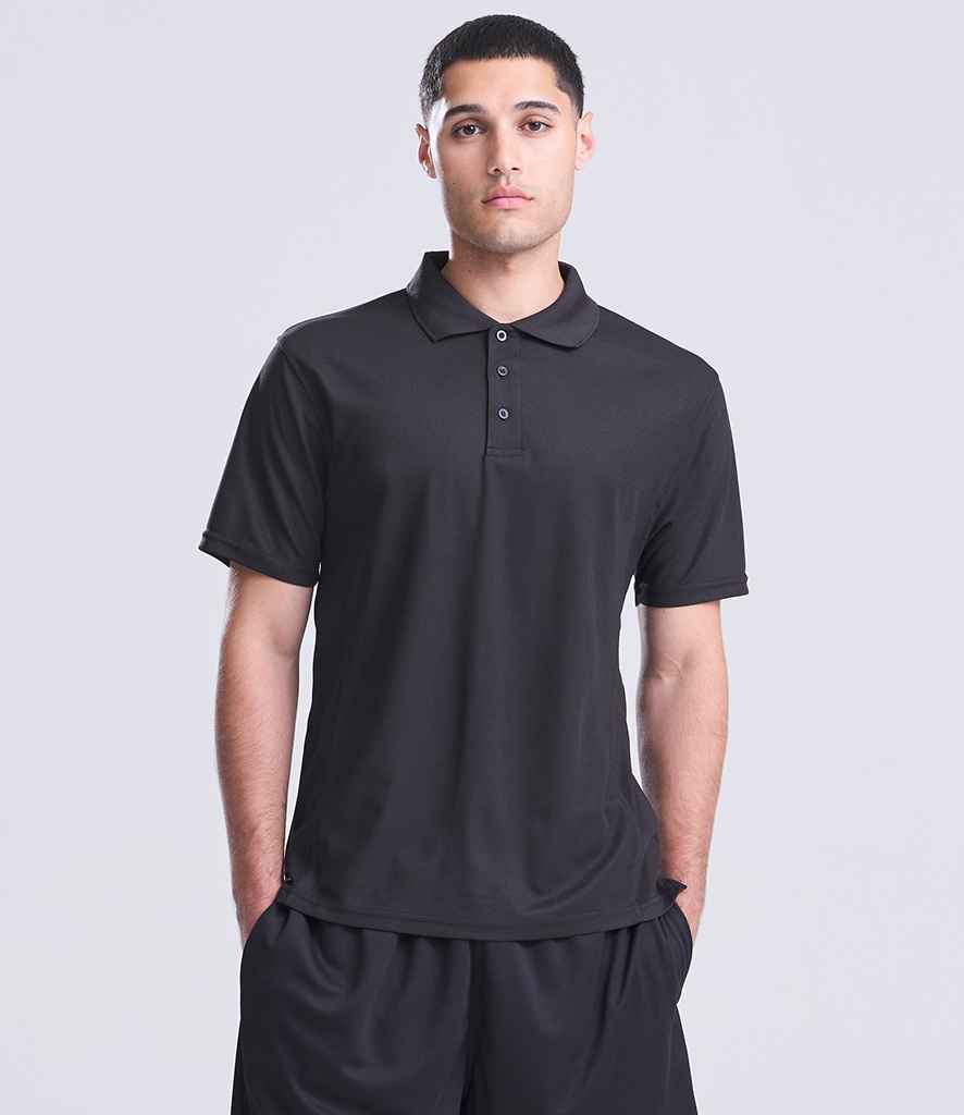 A person stands facing forward, wearing a Just Cool AWDis SuperCool™ Performance Polo Shirt in black with black shorts, hands in their pockets against a neutral light gray background.