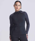 A woman faces forward wearing a black AWDis Ladies Cool Contrast Zoodie by Just Cool, featuring dark gray sleeves, front pockets, thumbholes on the cuffs, a zip-up front, and drawstring hood. The background is plain light gray.