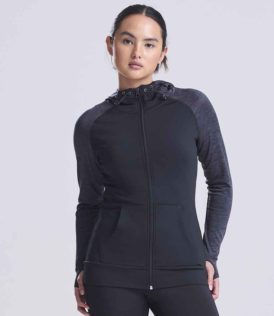 A woman faces forward wearing a black AWDis Ladies Cool Contrast Zoodie by Just Cool, featuring dark gray sleeves, front pockets, thumbholes on the cuffs, a zip-up front, and drawstring hood. The background is plain light gray.