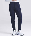 A person wears Just Cool AWDis Cool Tapered Jog Pants in navy blue with white athletic shoes, standing against a plain light background. Only the lower half of their body is visible.