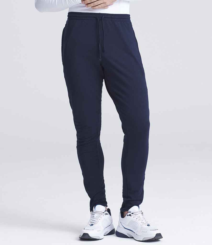 A person wears Just Cool AWDis Cool Tapered Jog Pants in navy blue with white athletic shoes, standing against a plain light background. Only the lower half of their body is visible.
