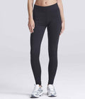 A person models AWDis Ladies Cool Athletic Pants by Just Cool—black, high-waisted leggings—paired with silver athletic sneakers against a plain light gray background. The upper body is cropped out of the image.
