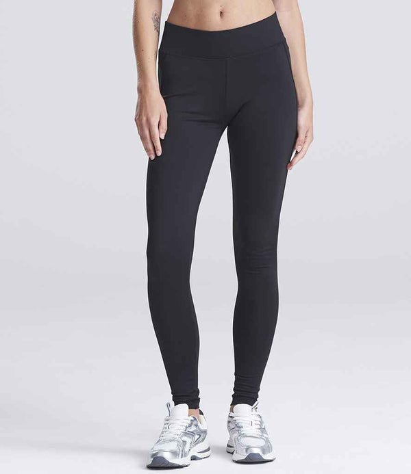 A person models AWDis Ladies Cool Athletic Pants by Just Cool—black, high-waisted leggings—paired with silver athletic sneakers against a plain light gray background. The upper body is cropped out of the image.