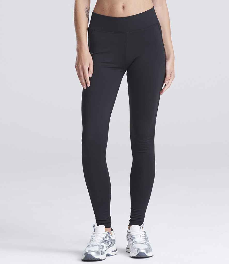 A person models AWDis Ladies Cool Athletic Pants by Just Cool—black, high-waisted leggings—paired with silver athletic sneakers against a plain light gray background. The upper body is cropped out of the image.