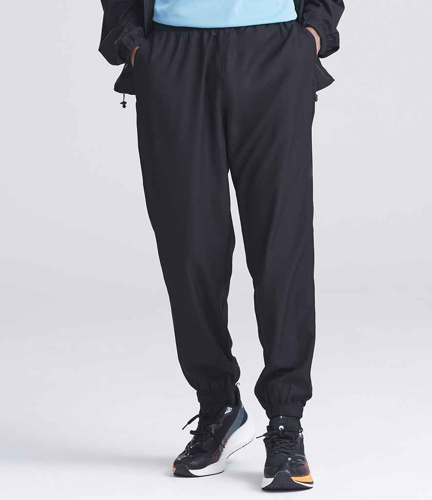 A person wearing Just Cool AWDis Cool Active Track Pants with zip pockets, a light blue shirt, and a black jacket stands with hands in pockets. The image, cropped at shoulders and feet, shows black athletic shoes with white soles.
