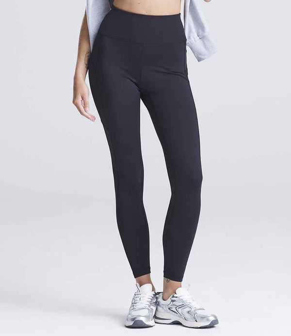 Wearing Just Cool AWDis Ladies Cool Recycled Tech Leggings in black, a gray top tied at the waist, and white athletic shoes, a person stands against a plain light background with their upper body partially out of frame.