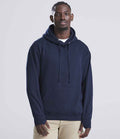 A man with short hair and a neutral expression faces the camera, wearing a navy blue AWDis Street Hoodie by Just Hoods over a white shirt and beige pants. The hoodie has a front pouch pocket, and the background is plain and light-colored.