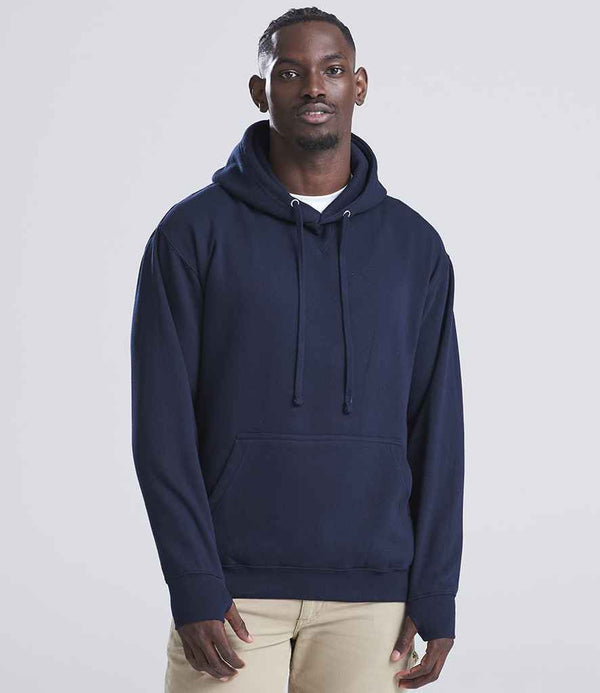 A man with short hair and a neutral expression faces the camera, wearing a navy blue AWDis Street Hoodie by Just Hoods over a white shirt and beige pants. The hoodie has a front pouch pocket, and the background is plain and light-colored.