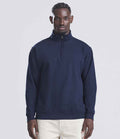 A man faces forward in a Just Hoods AWDis Sophomore Zip Neck Sweatshirt, navy blue with a YKK zip, paired with light pants against a plain light gray background.