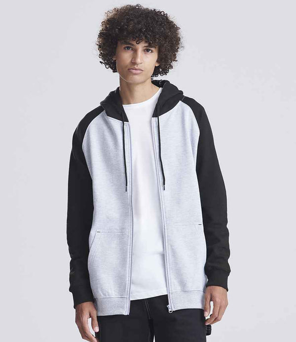 A person with curly hair is wearing the Just Hoods AWDis Baseball Zoodie, a light gray zip-up hoodie with black raglan sleeves, over a white t-shirt and standing against a plain light gray background.