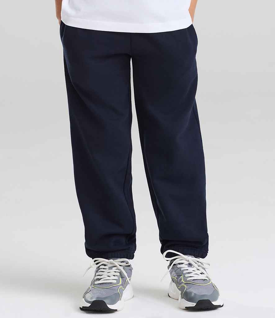 A person wears Just Hoods AWDis Kids Cuffed Jog Pants in navy blue with gray sneakers, standing against a plain light background. Only the lower half of the body is visible.