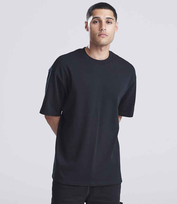 A person with short dark hair stands against a plain light background, wearing the Just Ts AWDis Signature Heavyweight T-Shirt in black and matching black pants, with their arms behind their back.
