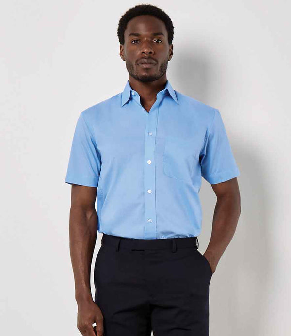 A man stands against a plain background wearing the Kustom Kit Short Sleeve Classic Fit Business Shirt by Kustom Kit in light blue, featuring a chest pocket and paired with dark trousers. One hand is in his pocket and his expression is neutral.