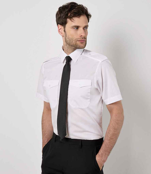 A man stands against a plain background wearing the Kustom Kit Short Sleeve Tailored Pilot Shirt, featuring chest pockets, paired with a black tie and pants. His hands are in his pockets and he gazes to the side.