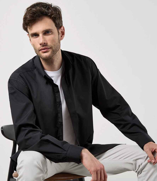 A man with short brown hair and light facial hair wears a Kustom Kit Long Sleeve Classic Fit Workforce Shirt in black over a white t-shirt with light pants, seated on a chair and looking at the camera against a plain background.
