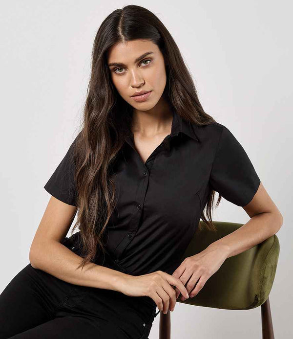 A woman with long brown hair wears a Kustom Kit Ladies Short Sleeve Tailored Poplin Shirt by Kustom Kit and black pants, sitting on a green chair against a plain light background, looking directly at the camera.