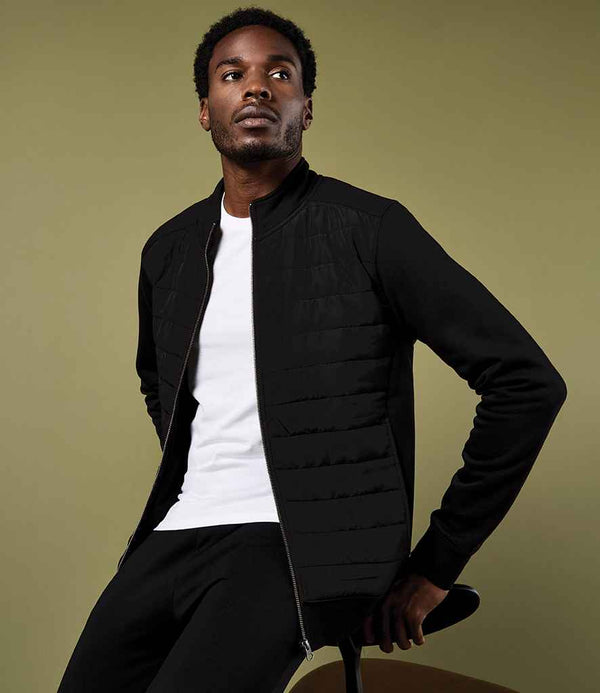 A man wearing the Kustom Kit Regular Fit Quilted Full Zip Sweat Jacket over a white t-shirt and black pants sits on a stool against an olive-green background, looking slightly upward and to the left.