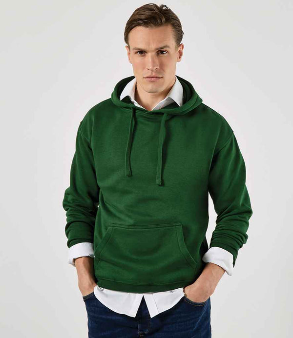 A person wears the Kustom Kit Hoodie by Kustom Kit—a dark green, drop-shoulder style with a jersey-lined hood—over a white collared shirt and dark jeans, hands casually in the front pouch pocket.