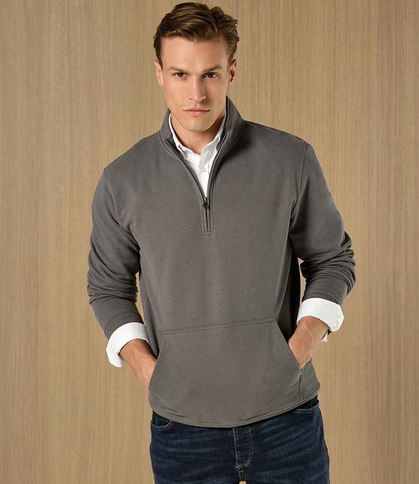 A person stands against a wood-textured background wearing a Kustom Kit Regular Fit 1/4 Zip Piqué Sweatshirt in gray over a white collared shirt and blue jeans, with their hands in the pullovers front pocket.