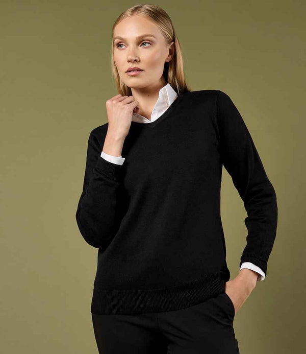 A person with long blonde hair wears the Kustom Kit Ladies Arundel Cotton Acrylic V Neck Sweater over a white collared shirt and black pants, standing against an olive green background with one hand touching their chin.