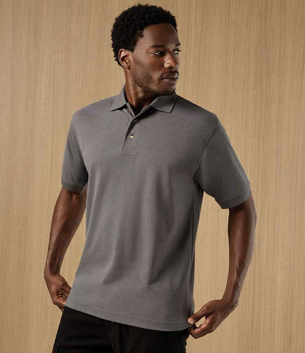 A man wearing the Kustom Kit Workwear Piqué Polo Shirt in gray stands with his arms relaxed by his sides, looking to his left in front of a light wooden background.
