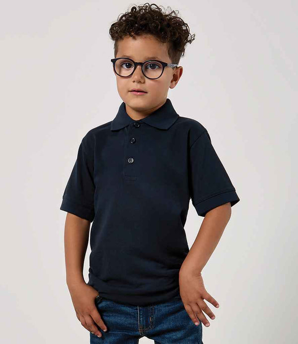 A young child with curly hair and glasses wears a Kustom Kit Kids Klassic Poly/Cotton Piqué Polo Shirt in dark navy, featuring a self-colour button placket and ribbed knit collar, paired with blue jeans, against a light background.