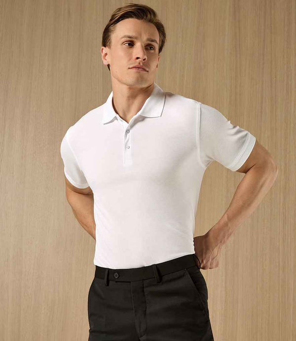 A man stands in front of a light wood panel, hands on hips, wearing the Kustom Kit Klassic Slim Fit Poly/Cotton Piqué Polo Shirt—a fitted white short-sleeve polo with pearlised buttons and a ribbed knit collar—from Kustom Kit.