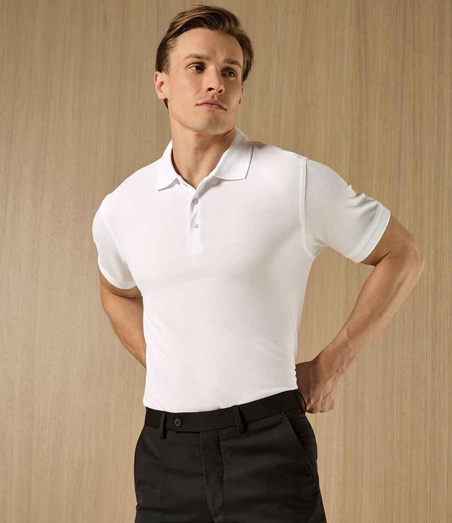 A man stands in front of a light wood panel, hands on hips, wearing the Kustom Kit Klassic Slim Fit Poly/Cotton Piqué Polo Shirt—a fitted white short-sleeve polo with pearlised buttons and a ribbed knit collar—from Kustom Kit.