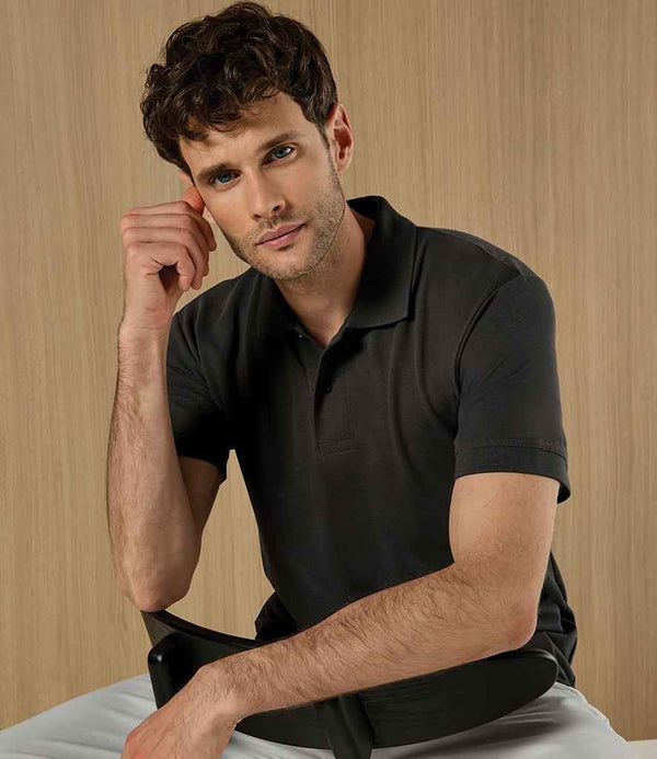 A man with short, curly dark hair and light skin wears a black Kustom Kit Regular Fit Recycled Superwash® 60° Polo Shirt and light pants. He sits on a chair against a light wooden background, resting his left elbow on his knee and touching his face.