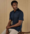A man in a Kustom Kit Regular Fit Cooltex® Plus Superwash® 60°C Piqué Polo Shirt (dark navy) and light beige pants sits on a wooden chair before a light wood-paneled background, looking slightly to his left.
