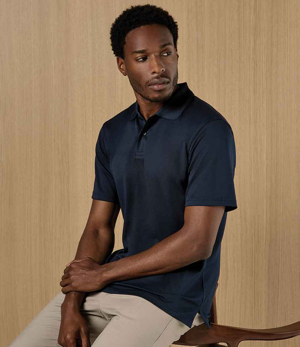 A man in a Kustom Kit Regular Fit Cooltex® Plus Superwash® 60°C Piqué Polo Shirt (dark navy) and light beige pants sits on a wooden chair before a light wood-paneled background, looking slightly to his left.