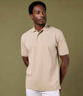 A man in a beige Kustom Kit Cotton Klassic Superwash® 60°C Polo Shirt with a ribbed collar and white pants stands against an olive-green background, gazing slightly left with his hands behind his back.